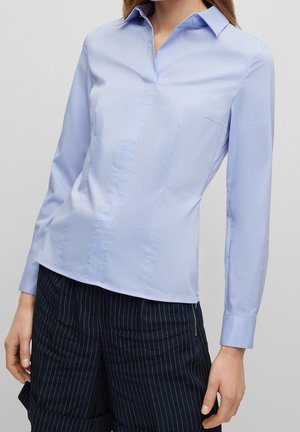 Femme portant un chemisier bleu clair à manches longues avec col et un pantalon noir à fines rayures, debout avec une main détendue le long du corps.