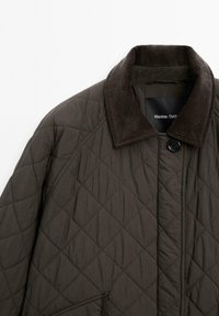 Kurtka o pikowanej fakturze w ciemnooliwkowym kolorze z kołnierzem z sztruksu; posiada zamek na guziki oraz diagonalny wzór przeszyć. Minimalistyczny design bez widocznych kieszeni.