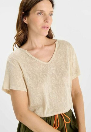 Femme aux cheveux châtain clair portant un haut beige à manches courtes et col en V, et un pantalon vert foncé avec un cordon orange, regardant vers la droite.