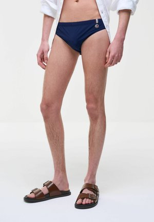 Uomo che indossa slip blu navy, camicia bianca sbottonata e sandali marroni con fibbia, in piedi davanti a uno sfondo bianco semplice.