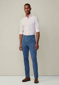 Άνδρας φορώντας ένα ανοιχτό ροζ πουκάμισο με rolled sleeves, μπλε παντελόνι προσαρμοσμένο και καφέ κομψές παπουτσές, στέκεται σε έναν απλό φόντο.