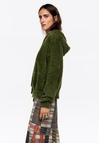 Sudadera verde y peluda con capucha. La modelo lleva una falda de cuadros con tonos rojos y grises. Mangas ajustadas, forma relajada.