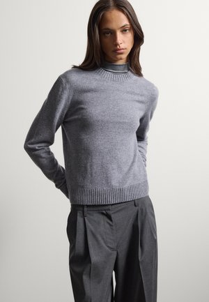 Joven con cabello largo y castaño, que lleva un jersey gris sobre un cuello alto y pantalones plisados gris oscuro, de pie frente a un fondo sencillo y claro.