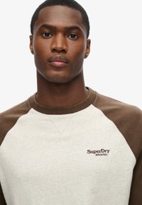 Bluza crewneck w szarym kolorze z brązowymi rękawami raglanowymi. Z przodu wyszywane logo "SuperDry Athletics". Miękka struktura materiału.