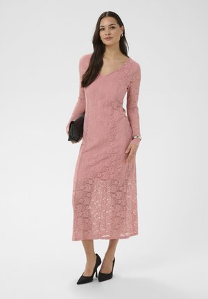 Kobieta w różowej koronkowej sukience midi z długim rękawem, czarnych szpilkach, trzymająca czarną kopertówkę, stojąca na białym tle.