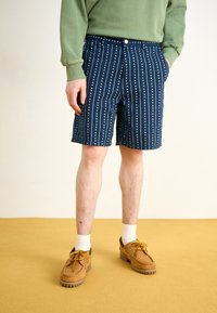 Marineblå shorts med vertikale hvite prikker, laget av et lett stoff, med knappelukking og sidelommer, kombinert med brune sko.