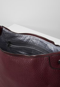 Sac à main en cuir bordeaux avec une finition texturée, doté d'une fermeture éclair et d'un intérieur à motifs gris avec une petite poche zippée.