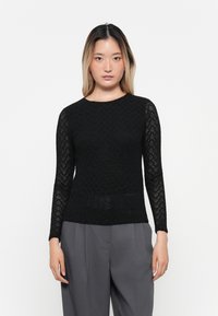 Black long-sleeve top with a textured lace pattern, featuring a round neckline and fitted silhouette, paired with gray trousers.