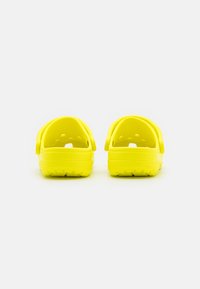 Une paire de sabots jaunes vifs à enfiler avec des brides au dos, vue de l'arrière sur un fond blanc.