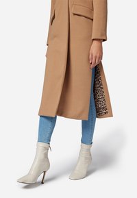 Cappotto lungo beige e strutturato con fodera leopardata. Indossato su jeans azzurri chiari e stivaletti bianchi con punta affusolata e tacco a stiletto.