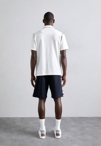 Λευκό πόλο μπλουζάκι με μοτίβο στους ώμους, σε συνδυασμό με μπλε σορτς. Λευκές κάλτσες και αθλητικά παπούτσια με ζικ-ζακ μοτίβο ολοκληρώνουν την εμφάνιση.