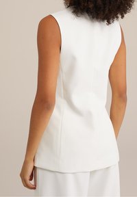 Ärmlös vit skräddarsydd topp bärs av en person med lockigt hår, visad från baksidan mot en enfärgad beige bakgrund.