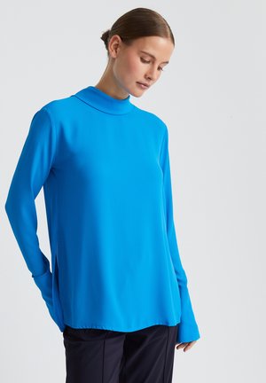 Langarm turtleneck top in levendig blauw. Glad materiaal met een relaxed fit, zijsplitsingen aan de zoom en verlengde mouwen. Neutrale achtergrond.