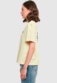 Gul bomull T-skjorte med korte ermer, avslappet passform og logo trykk på siden. Brukes sammen med lyseblå denimjeans, stående mot en nøytral bakgrunn.