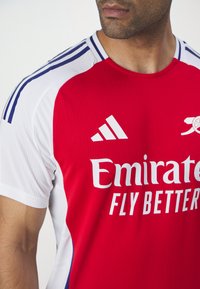 Maillot de sport rouge et blanc à manches courtes, présentant un tissu texturé, des accents bleus sur les épaules et les logos d'Adidas et d'Emirates.