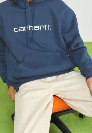 Osoba ubrana w granatową bluzę z kapturem Carhartt i kremowe spodnie, siedząca na pomarańczowym fotelu biurowym na zielonej podłodze.