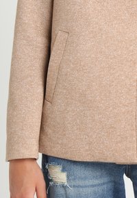 Beige tekstureret frakkeærme og lomme med en hånd ved siden af slidte blå denim jeans.