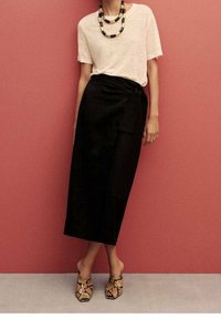 Jupe noire enveloppante assortie à un haut beige à manches courtes, portant un collier en perles multicolores et des chaussures en imprimé serpent. Textures de tissu lisses visibles.