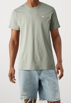 Lysegrøn bomulds-T-shirt med rund hals og korte ærmer, med en broderet drikkeglas på den øverste venstre bryst. Bær med slidte denimshorts.