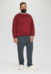Bearded man in een rode trui, donkergrijze broek en witte sneakers, staand tegen een effen lichte achtergrond.