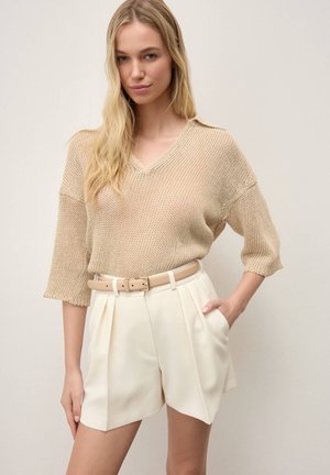 Blondīne sieviete, kas valkā bēša krāsas adītu V-veida kakla džemperi ar elkoņa garuma piedurknēm, krēmkrāsas augsto vidukli kroku šorti un bēša krāsas jostu, roka kabatā.