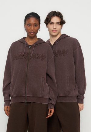 Két modell páros barna cipzáras kapucnis pulóvert visel, melyeken texturált "VISION" felirat található, és egy egyszerű világos háttér előtt állnak.