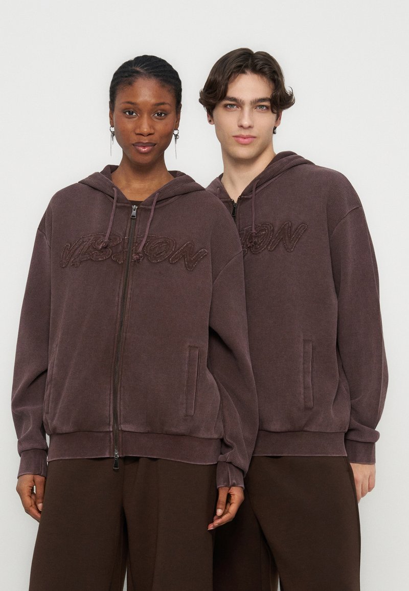 Δύο μοντέλα φοράνε ταιριαστά καφέ φούτερ με φερμουάρ και κείμενο "VISION" με υφή, στέκονται μπροστά σε απλό φωτεινό φόντο.