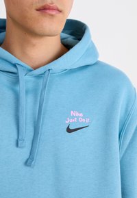 Vaaleansininen huppari, joka on valmistettu pehmeästä kankaasta ja jossa on pinkki-musta "Nike Just Do It" -logo rinnassa sekä kiristysnauhallinen huppu.