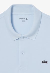 Ljusblå, texturerad polo tröja tillverkad av en blandning av bomull och polyester. Har en treknappslås och broderad Lacoste-logga på bröstet.
