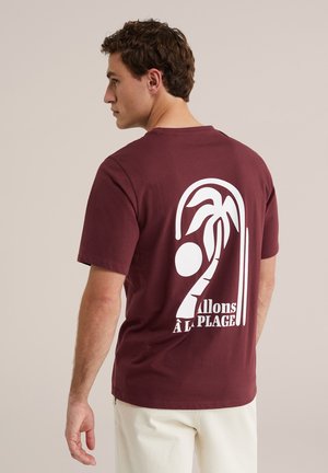 Homme aux cheveux courts et bruns portant un t-shirt bordeaux avec un motif palmier blanc et un texte en français, associé à un pantalon clair.