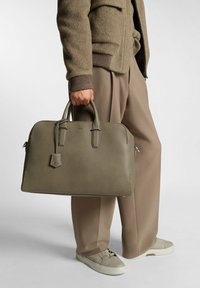 Leather handbag in muted green, rectangular shape with two top handles, zip closure, and luggage tag detail; textured finish.