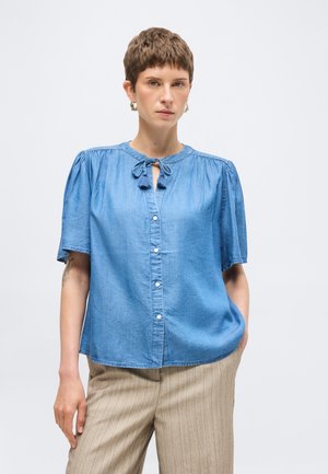 Frau mit kurzen Haaren, die goldene Creolen, eine blaue kurzärmlige Bluse mit Knöpfen und Bindeband am Ausschnitt sowie beige gestreifte Hose trägt.
