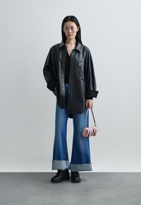 Black leather oversized shirt jacket over a black top, paired with wide-leg denim jeans featuring frayed hems and black platform shoes. Holds a small pink bag.