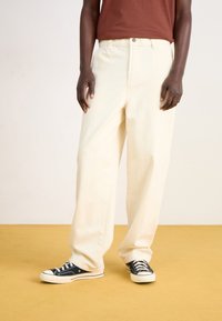 Pantalon en denim crème à jambes larges, doté de poches avant, d'une fermeture à bouton, associé à des baskets basses noires.