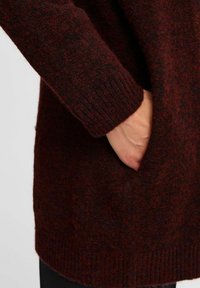 Maglione bordeaux lavorato a maglia con tessuto testurizzato, polsini a coste e una tasca laterale. Materiale soffice con una leggera lucentezza e variazioni di colore sottili.