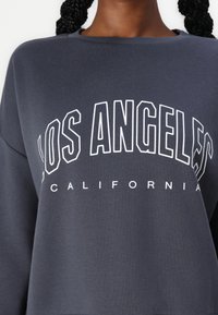 Mørkegrå sweatshirt med "LOS ANGELES" i store hvide konturerede bogstaver og "CALIFORNIA" i mindre hvide bogstaver nedenunder.