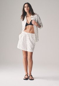 Weißes Hemd- und Shorts-Set mit schwarzen Polka-Dots, gefertigt aus leichtem Stoff. Inklusive schwarzem Bikini-Top und schwarzen Sandalen.