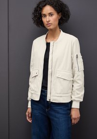 Veste bomber beige clair avec texture côtelée, fermeture éclair, poches latérales, et une poche zippée supplémentaire sur la manche. Associée à un jean foncé.