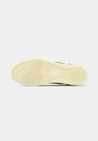 Semelle de chaussure orientée vers le haut montrant une semelle extérieure en caoutchouc beige avec des motifs en chevrons et circulaires sur fond blanc.