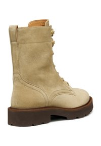 Stivale alla caviglia in suede beige con punta arrotondata, cuciture dettagliate, hardware in ottone e una spessa suola in gomma marrone. Caratterizzato da lacci sulla parte anteriore e anello sul retro.