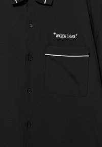 Chemise noire boutonnée avec une poche poitrine bordée d'un liseré blanc et un texte brodé en blanc "WATER SIGNS" avec des accents d'étoiles au-dessus.