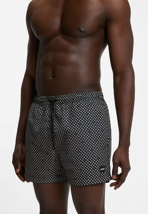 Skjorteløs mand iført sorte badeshorts med hvidt og beige geometrisk stjernemønster og sort snøre, stående mod en hvid baggrund.