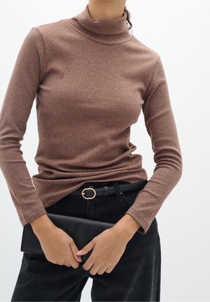Personne portant un pull à col roulé marron côtelé et un pantalon noir, tenant une pochette noire rectangulaire devant un fond blanc uni.