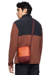 Borsa a tracolla arancione e rossa con texture liscia, forma rettangolare e tracolla regolabile, indossata sopra una giacca in pile marrone e nera.