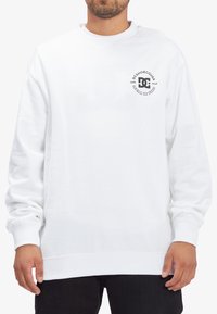 White cotton sweatshirt with long sleeves, round neck, and a circular black logo on the left chest. Features ribbed cuffs and hem.