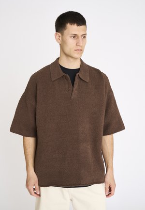 Mand iført en løs, brun tekstureret kortærmet skjorte med krave og knapper, stående mod en ensartet hvid baggrund.