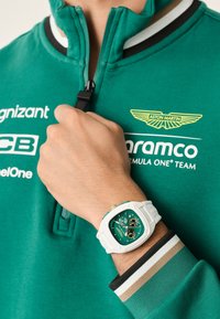 Fehér sportóra négyzet alakú tokra, zöld számlapra és fekete alacsony szíjakra. Egy kézen viselve, zöld zipzáros pulóverrel párosítva, amely csíkos.