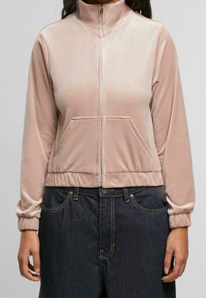 Femme portant une veste zippée en velours rose clair avec une poche avant et un jean taille haute foncé, debout devant un fond uni.