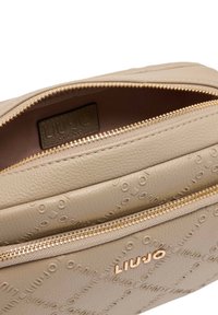 Bolso de cuero beige texturizado con herrajes dorados, que cuenta con una cremallera superior y un logo en relieve, revelando un interior espacioso.