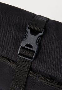 Zwarte plastic zijklepbeugel die een verstelbare zwarte nylon strap op een tas van textuurstof beveiligt.
